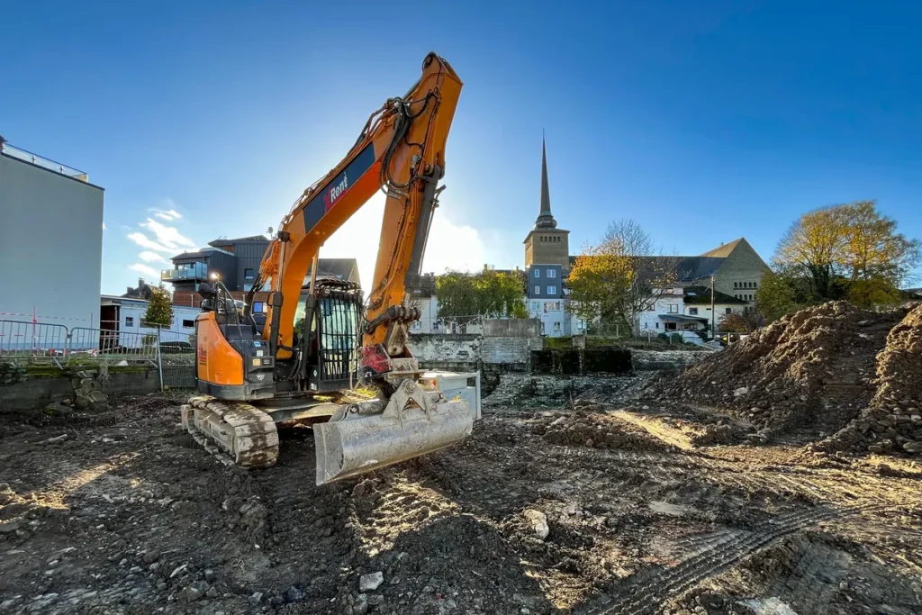 Orangefarbener Bagger auf dem Grabungsgelände in St. Vith, dahinter ein Kirchturm