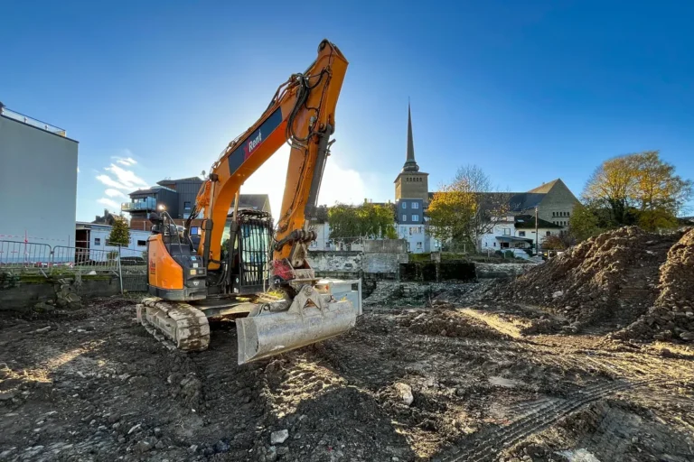 Orangefarbener Bagger auf dem Grabungsgelände in St. Vith, dahinter ein Kirchturm