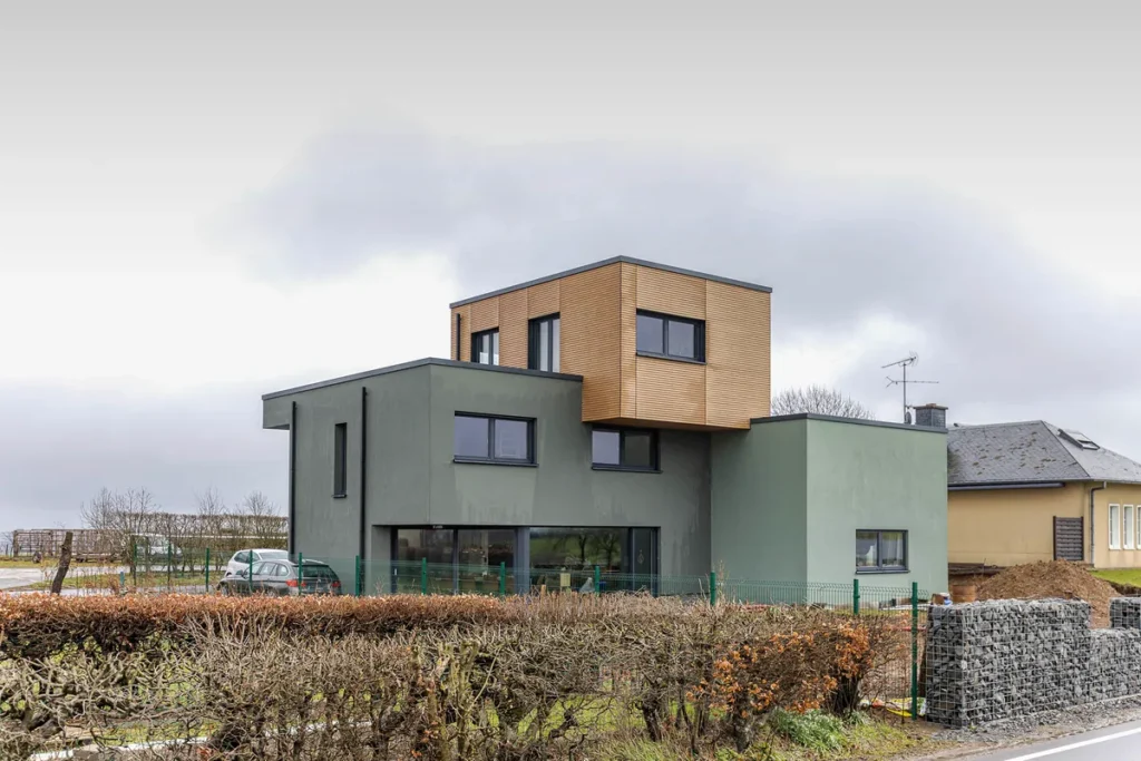 Kubisches Einfamilienhaus mit grüner Fassade und braun verkleidetem Obergeschoss, davor Hecke